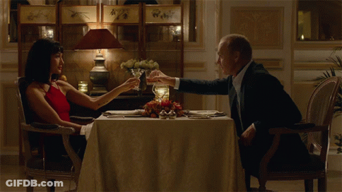 A Man And Woman Are Sitting At A Table With Glasses Of Wine GIF