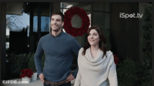 A Man And Woman Are Standing In Front Of A Building With A Wreath In The Background And Ispot.tv On The Bottom Right GIF