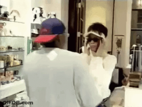 A Man And Woman Are Standing In Front Of A Mirror In A Room . The Woman Is Wearing Sunglasses And A Hat . GIF