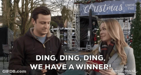 A Man And Woman Are Standing In Front Of A Sign That Says Christmas Under The Christmas Tree GIF