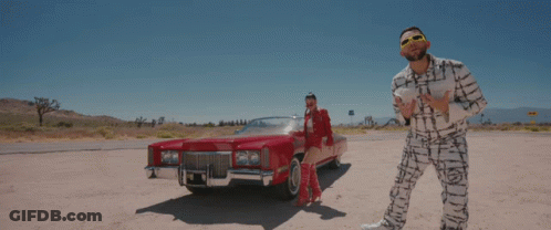 A Man And Woman Are Standing Next To A Red Car GIF