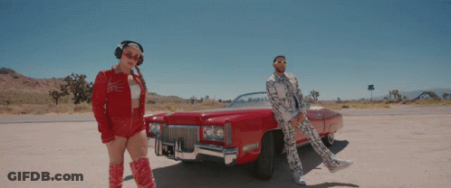 A Man And Woman Are Standing Next To A Red Car GIF