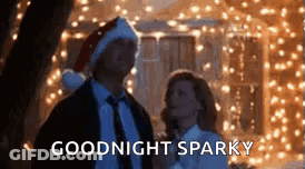 A Man And Woman Are Standing Next To Each Other In Front Of Christmas Lights And The Man Is Wearing A Santa Hat . GIF