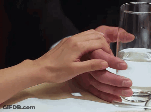 A Man And Woman Holding Hands Over A Glass Of Water On A Table GIF