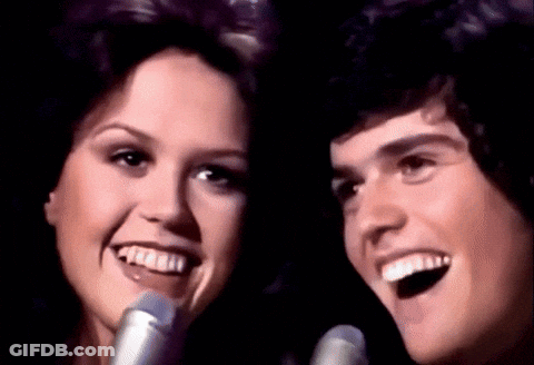 A Man And Woman Singing Into Microphones And Smiling GIF