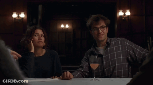 A Man And Woman Sitting At A Table With A Glass Of Wine GIF