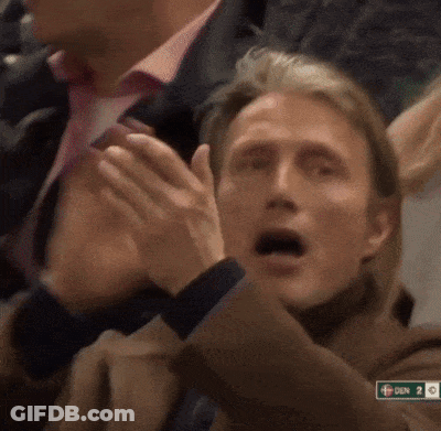 A Man Applauds While Sitting In A Crowd With A Sign That Says Den On It GIF