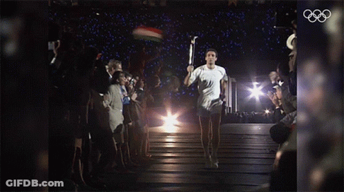 A Man Carrying A Torch In Front Of A Crowd With The Olympic Rings In The Corner GIF