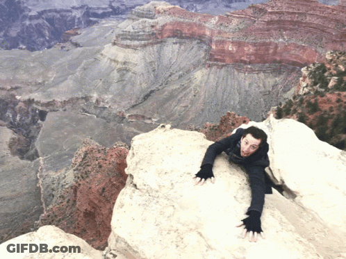 A Man Climbs Up A Rocky Cliff With A Valley In The Background GIF