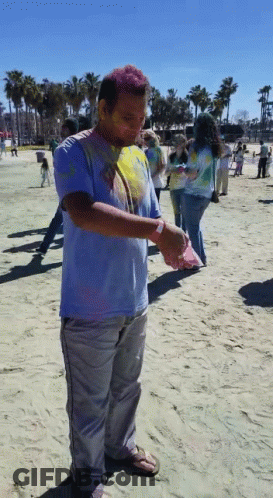 A Man Covered In Colored Powder Stands On A Sandy Beach GIF