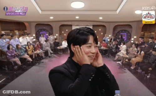 A Man Covering His Ears With His Hands In Front Of A Crowd With A Kbs Logo On The Ceiling GIF