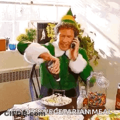 A Man Dressed As A Christmas Elf Is Talking On A Cell Phone While Sitting At A Table With Food . GIF