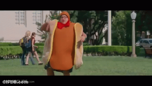 A Man Dressed As A Hot Dog Is Walking In A Park . GIF