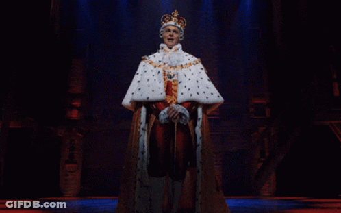 A Man Dressed As A King With A Crown On His Head Stands On A Stage GIF