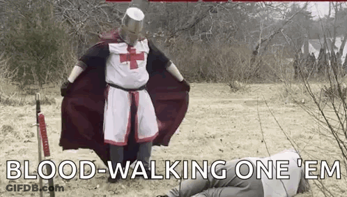 A Man Dressed As A Knights Templar Is Standing Next To A Man Laying On The Ground With A Sword . GIF