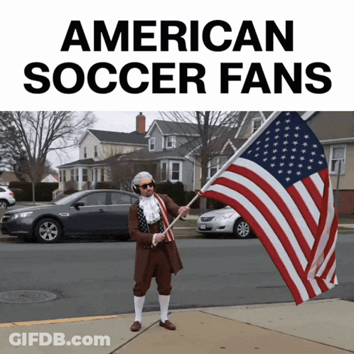 A Man Dressed As Benjamin Franklin Holds An American Flag In Front Of A Sign That Says American Soccer Fans GIF