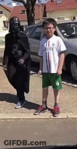 A Man Dressed As Darth Vader Stands Next To A Young Boy Wearing A White Shirt That Says Serie GIF