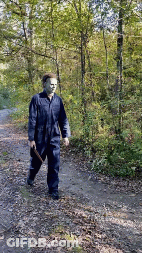 A Man Dressed As Michael Myers Is Walking Down A Path In The Woods GIF