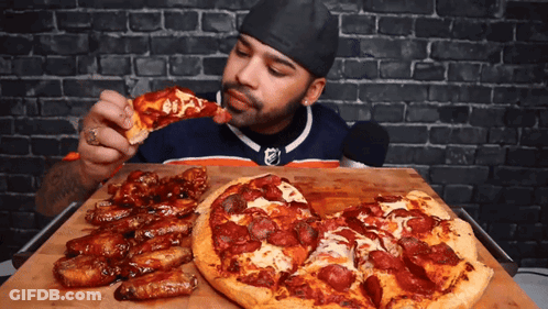 A Man Eating A Slice Of Pizza Next To A Pizza With Pepperoni On It GIF