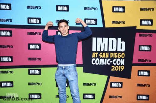 A Man Flexes His Muscles In Front Of A Colorful Backdrop That Says Imdb At San Diego Comic Con 2019 GIF