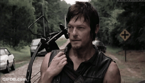A Man Holding A Bow And Arrow Stands In Front Of A Road Sign That Says Peace Love And Curves GIF