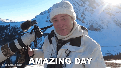 A Man Holding A Camera In Front Of A Snowy Mountain With The Words Amazing Day Above Him GIF