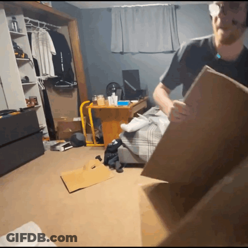 A Man Holding A Cardboard Box In A Messy Room With A Shirt That Says " I Love You " GIF