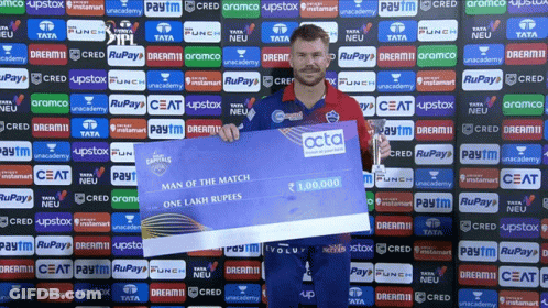 A Man Holding A Cheque In Front Of A Wall With Advertisements For Tata And Ceat GIF