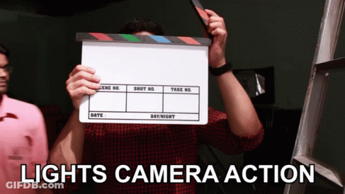 A Man Holding A Clapper Board With The Words Lights Camera Action GIF