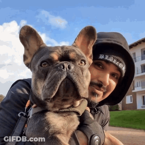 A Man Holding A Dog Wearing A Black Beanie That Says Blaze GIF