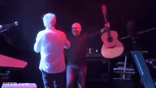 A Man Holding A Guitar On Stage With Another Man GIF
