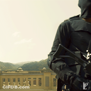 A Man Holding A Gun In Front Of A Building And Mountains GIF