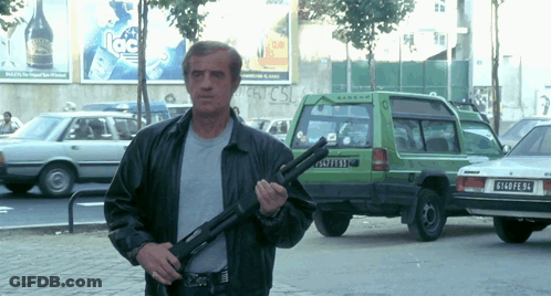 A Man Holding A Gun In Front Of A Green Car With The License Plate R140pe94 GIF