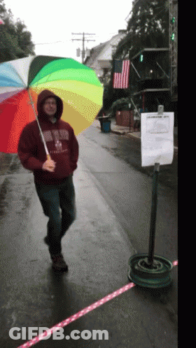 A Man Holding A Rainbow Colored Umbrella Is Wearing A Sweatshirt That Says ' Boston ' On The Front GIF
