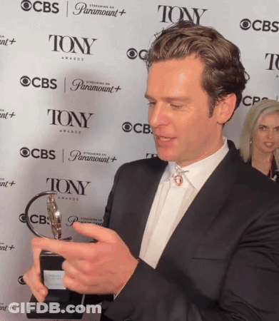 A Man Holding A Trophy In Front Of A Wall That Says Tony Awards GIF