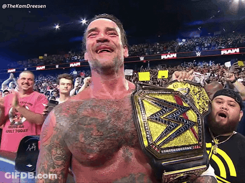 A Man Holding A Wrestling Championship Belt In Front Of A Crowd With A Sign That Says Raw GIF