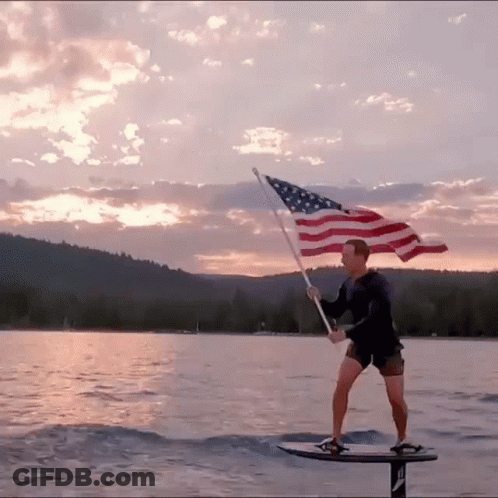 A Man Holding An American Flag While Standing On A Surfboard GIF