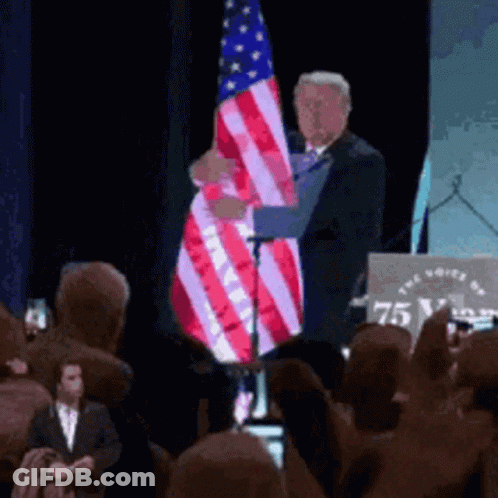 A Man Holds An American Flag In Front Of A Podium That Says 75 GIF