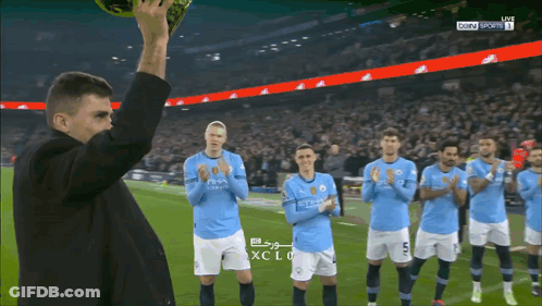 A Man Holds Up A Soccer Ball In Front Of A Group Of Soccer Players Wearing Blue Shirts With The Word City On Them GIF