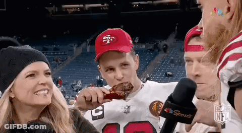 A Man In A 49Ers Hat Is Eating A Chicken Wing At A Football Game While A Woman Looks On . GIF