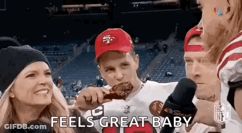 A Man In A 49Ers Hat Is Eating A Piece Of Meat While A Woman Holds A Child . GIF
