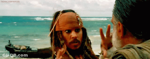 A Man In A Bandana Is Talking To Another Man On A Beach With A Boat In The Background . GIF