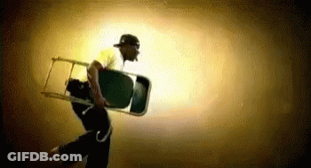A Man In A Baseball Cap Is Carrying A Chair In A Dark Room . GIF