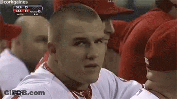 A Man In A Baseball Uniform Is Sitting In A Dugout Looking At The Camera . GIF