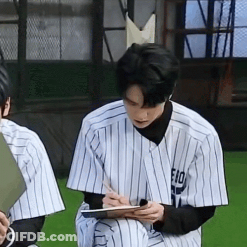 A Man In A Baseball Uniform Is Sitting On The Grass Writing On A Clipboard . GIF