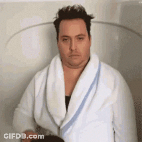A Man In A Bathrobe Is Sitting In A Bathtub With A Towel Around His Neck . GIF
