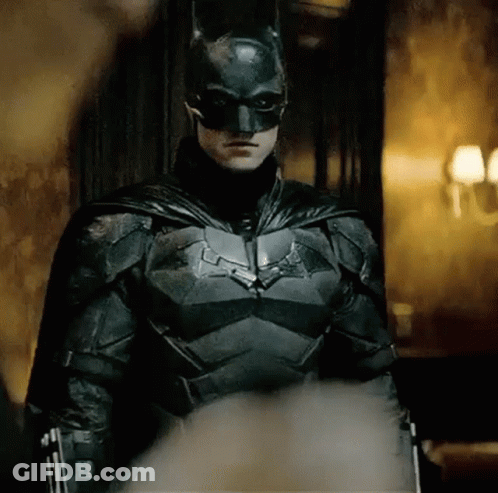 A Man In A Batman Costume Is Standing In A Room With A Gun . GIF