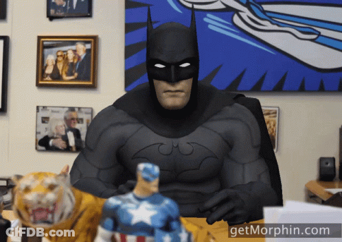A Man In A Batman Costume Sits At A Desk With A Tiger And A Captain America Figurine GIF