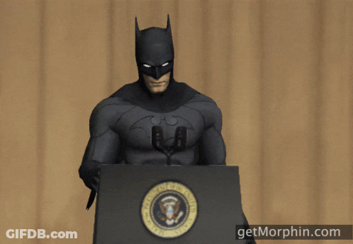 A Man In A Batman Costume Stands At A Podium With A Microphone In His Hand GIF