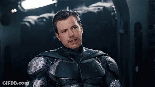 A Man In A Batman Suit Is Sitting In A Dark Room And Pointing At The Camera . GIF